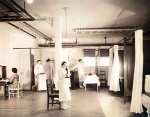 A treatment room. Nurses in white dresses attend to patients in beds behind white curtains.