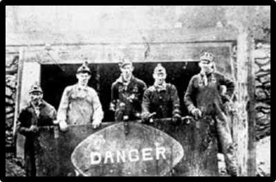 Group of miners wearing hard hats at the entrance of a shaft, standing behind a sign that reads: DANGER.