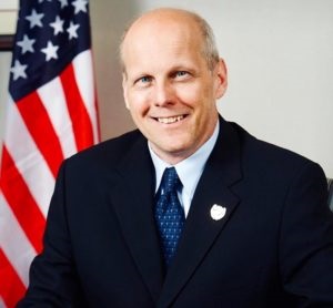 Commissioner Krickus in a dark blazer, blue tie, in front of an American flag.