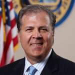 Commissioner Mastrangelo, in a dark blazer and blue tie, in front of an American flag