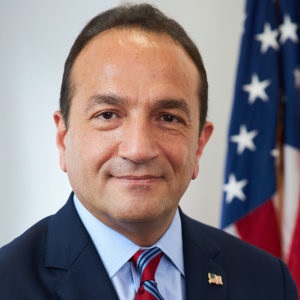 Commissioner Selen in a blue suit and red tie, standing in front of an American flag