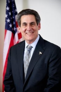 Commissioner Shaw in a dark suit and light gray tie, in front of an American flag