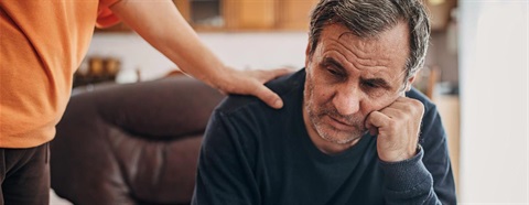 Older man looking sad, being comforted by someone standing above him