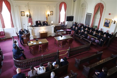 A courtroom with a red rug filled with judges and lawyers from Morris County. There is wooden furniture and two large windows behind a large and high court bench.
