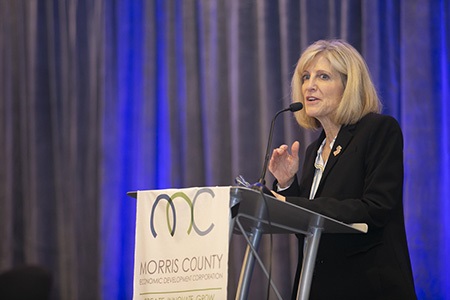 A woman,Christien Myers, speaking at a podium. She had short blonde hair and is wearing a dark suit.