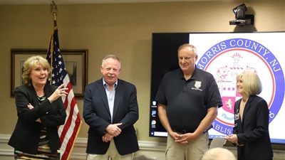 Two men stand in from of the Morris County logo, Lou Pepe and David Barter. On each side of them there is a woman, Cathy Burd and Commissioner Director Christine Myers. They are being honored upon their retirement. They also stand by an American flag.