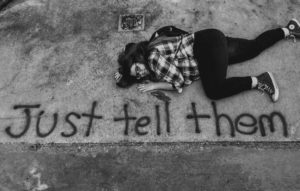 Black and white photo of a teenage girl lying on a concrete floor. At the bottom of the photo, the workds "Just Ttell them are spray painted..