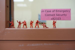 Photo of seven red plastic soldiers, children's toys, set up on an interior window ledge under a pink sign that says "In Case of Emergency, Contact Security" 