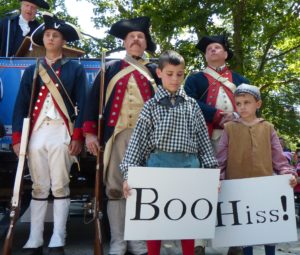 In period dress, soldiers stand behind children holding signs that say 'boo' and 'hiss.'