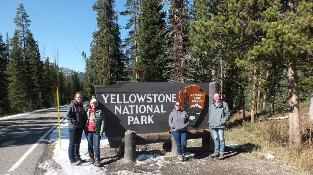 Four people standing in front of the Yellowstone National Park sign, which is on the side of a road. Some snow on the ground, lots of trees in the backgorund.